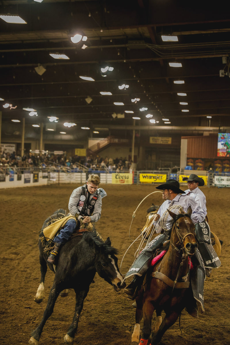 Pickup men on horseback ride alongside a saddle bronc rider under the lights of an indoor rodeo arena — the kind of night you dress for at the NFR in Las Vegas