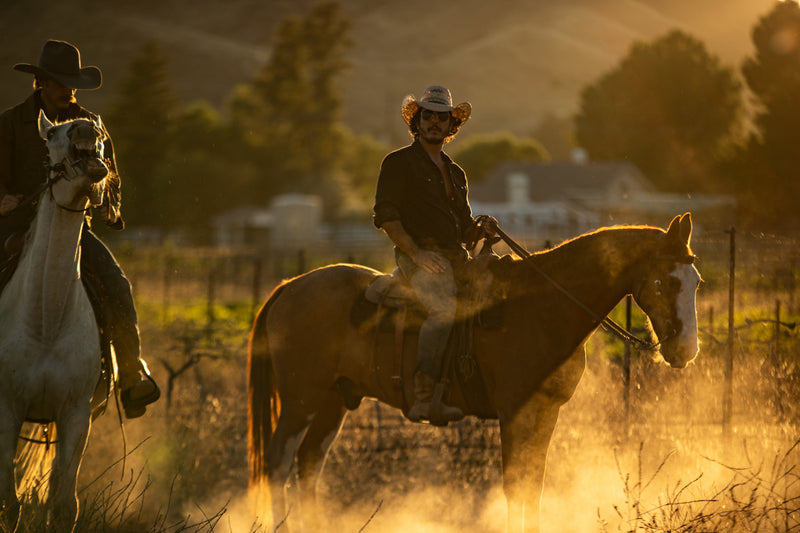 Two riders on horseback at golden hour with dust rising around the horses' legs -- Cowboy Church on riding drag and serving where nobody's looking