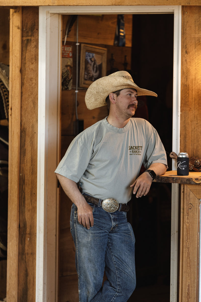 Cowboy in a straw cowboy hat and a Sackett Ranch tee leaning in the doorway of a wooden tack room with saddle pads behind him