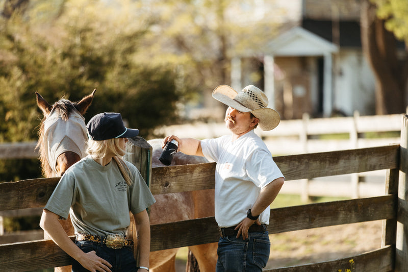 Man in a straw cowboy hat and a woman in a navy Sackett Ranch trucker cap talking at a wooden ranch fence beside a horse in golden evening light
