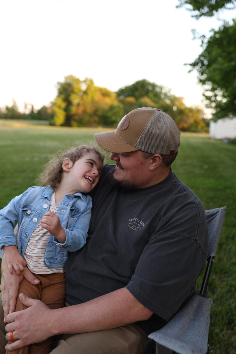 Smiling father in a tan Sackett Ranch trucker hat holding his laughing young daughter on his lap outdoors at golden hour