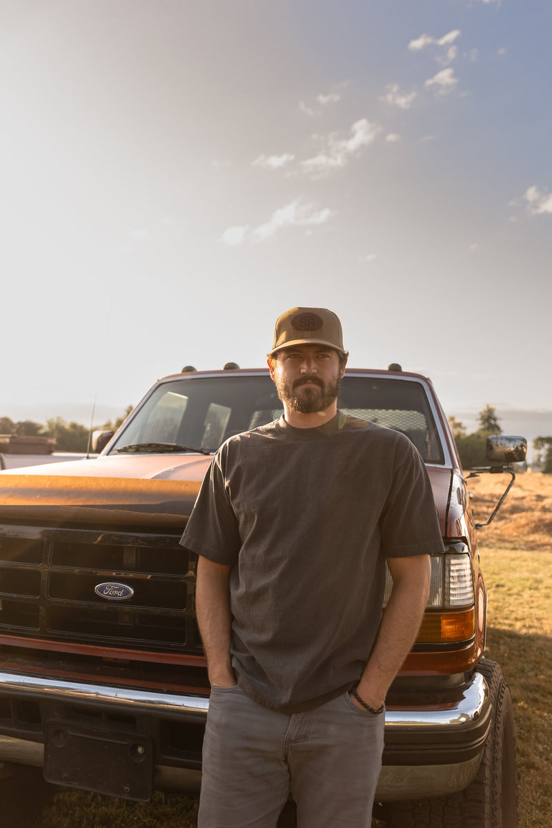 Bearded man in a faded black heavyweight cotton tee and a Sackett Ranch trucker hat standing in front of an old pickup at golden hour