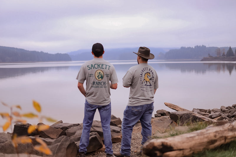 Two men in Sackett Ranch tees, one in a felt cowboy hat, standing on a rocky shore looking out over still water toward misty forested hills